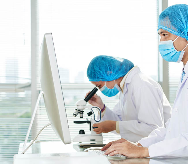 Team of talented microbiologists wearing white coats and medical masks wrapped up in work at modern lab: Asian woman studying sample with help of microscope while her male colleague taking notes
